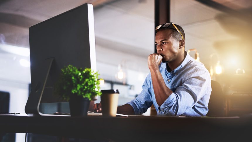 An African American man looking pensive stares at a large computer monitor in his professional office